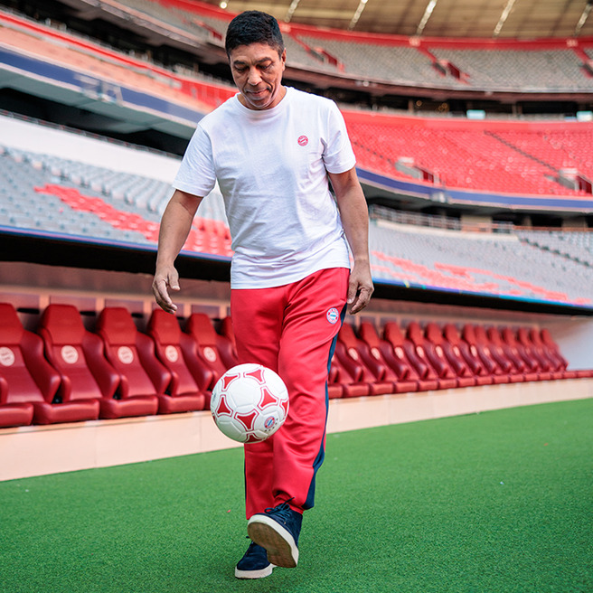 Futbol o‘ynayotgan odam, oq futbolka va qizil shalvar kiyib, to'pni ushlab turibdi. U keng, bo‘sh stadionda, o‘rindiqlar oldida joylashgan. बिल्कुल रोशनी में, u kulib, quvonch bilan to'pni tepadan tushiradi.