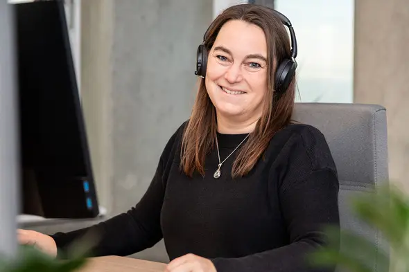 Ofisda kompyuter oldida o'tirgan, quloqchin taqilgan ayol, tabassum qilib nishonlayapti. Orqa fon shaffof va zamonaviy ko'rinishda.