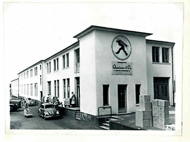 Qora va oq rangda eski binoni ko'rsatadigan foto, old tomonda logo va "Quinver & Co." yozuvi bor. O'rtada bir necha avtomobillar, tashqarida esa odamlar va qutichalar. Binoning ko'chalari va derazalarining ko'rinishi eski zamonga oid.