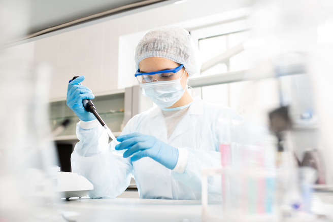 Laboratoriya sharoitida quyoshda ishlovchi kimyo mutaxassisi, pipetka bilan namuna olib, tajriba o'tkazmoqda. U himoya kiyimida, ko'zoynak va niqobda. Orqa fon hareketli, laboratoriya asboblari ko'rinadi.