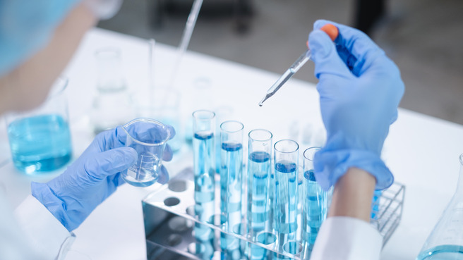 Laboratoriyada ko'k suyuqlik bilan to'ldirilgan test trubkalari ustida ishlovchi, ko'k himoya qo'lqog'i kiygan odam. U bir qo'lida pipetka, boshqa qo'lida esa kolba ushlab turadi.