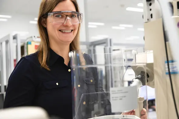 Laboratoriyada ishlayotgan ayol, himoya ko'zoynaklari bilan nafaqaga chiqqan qizil jihozdan foydalanmoqda, kulib turibdi. Fonida laboratoriya asboblari va ishlov berish joylari ko'rinadi.