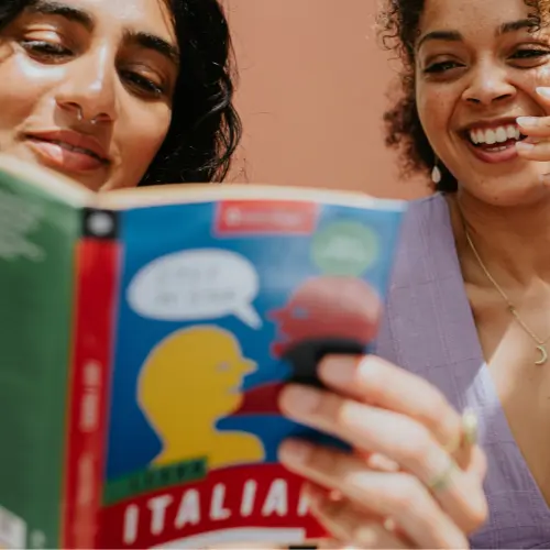 Две женщины читают книгу, улыбаются и обсуждают её. На книге яркая обложка с изображением двух фигурок и надписями. Фон стены розовый.