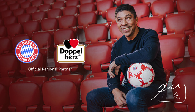 Bayern Munchen futbol klubi logotipi va Doppelherz brendi bilan birga, futbol to'pini ushlab turgan odatdagi stolda o'rnashgan odam. Odam tabassum qilib, ko'zlariga kuchli ishtiyoq bilan qaramoqda.
